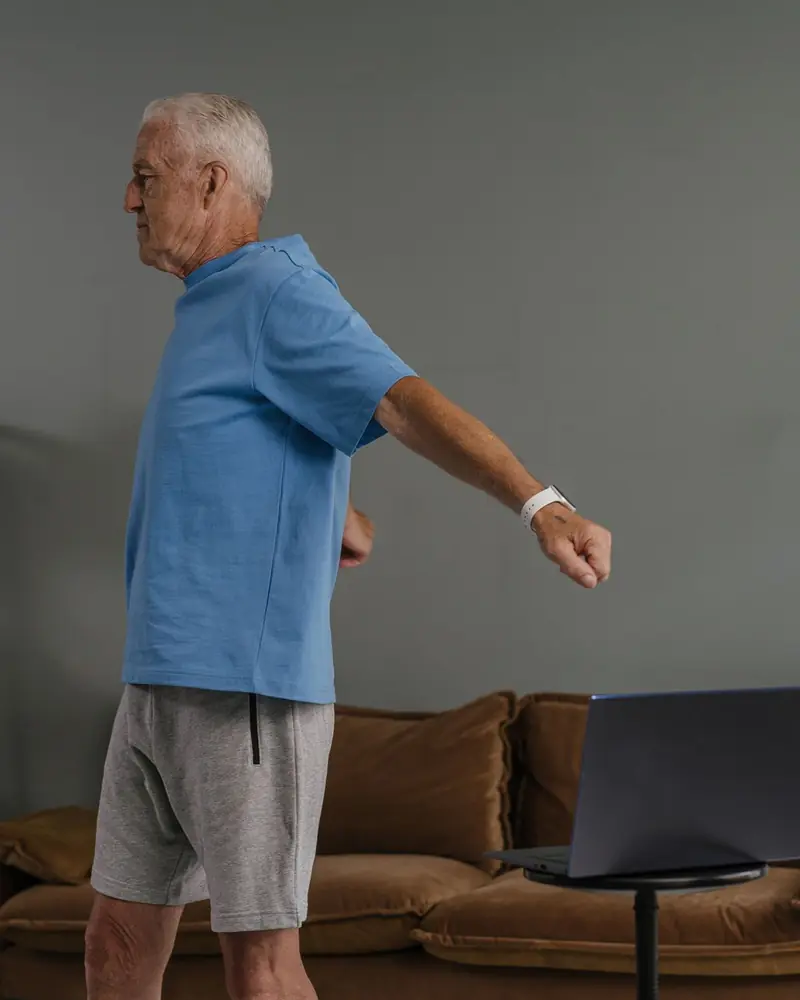 An older man doing a standing exercise at home, his session open on a laptop.