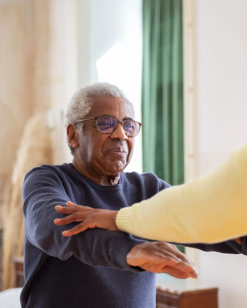 A seated patient working through arm movements at home while a helper guides the reach.