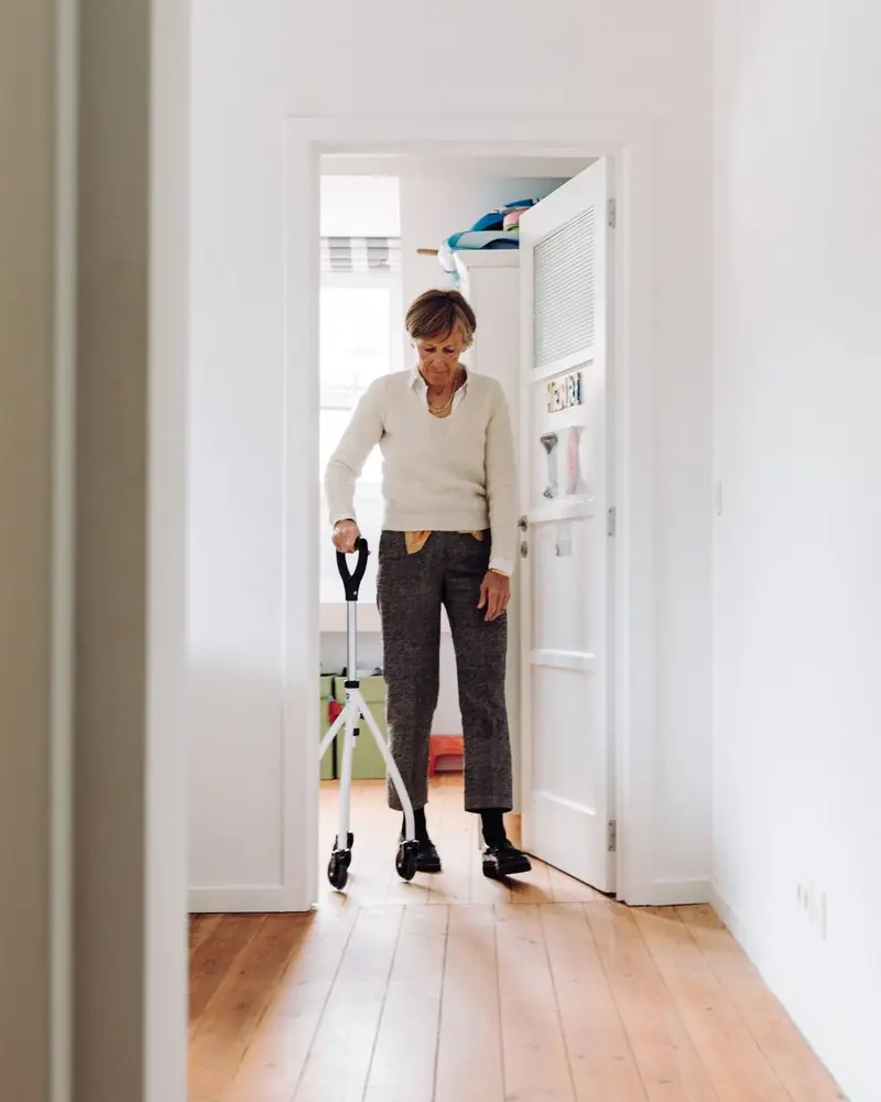 An older woman practising a deliberate step through a doorway at home, steadying on a walking aid.